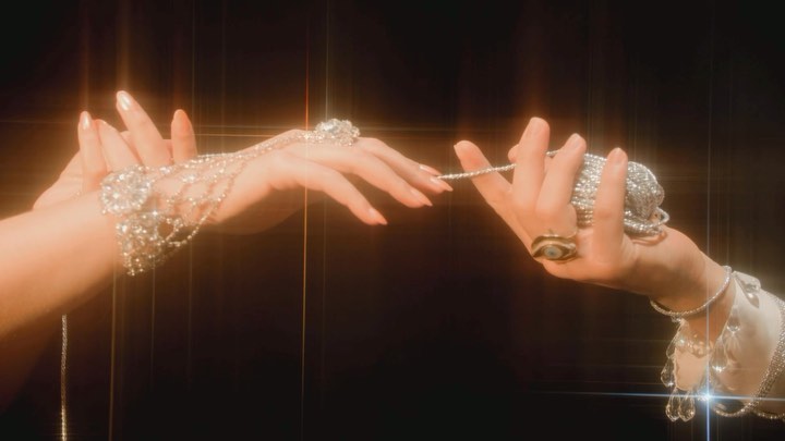 Close-up studio shot featuring two hands adorned with elaborate jewelry, including rings and bracelets, against a dark backdrop.