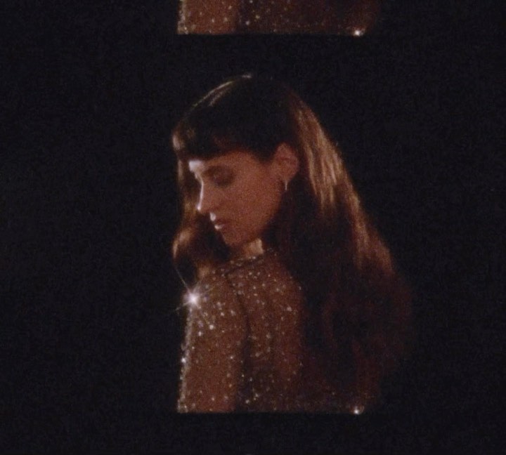 Portrait of a young adult woman with long brown hair and a sparkly top against a dark background in a dimly lit indoor setting.