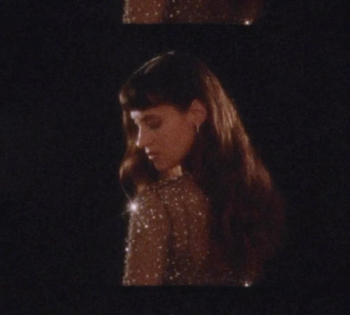 Portrait of a young adult woman with long brown hair and a sparkly top against a dark background in a dimly lit indoor setting.