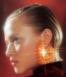 Close-up profile portrait of a woman wearing large orange jeweled earrings with a glowing starburst effect against a red backdrop.