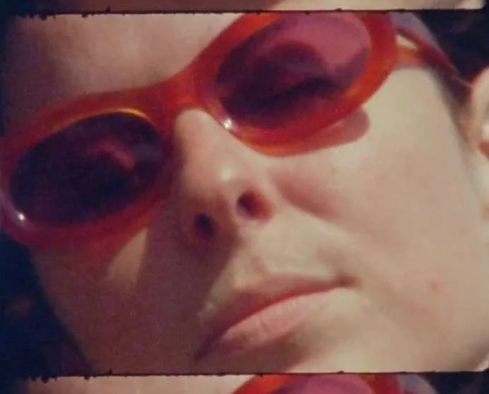 Close-up portrait of a person wearing vibrant orange-tinted sunglasses, captured on analogue film with visible sprocket holes.