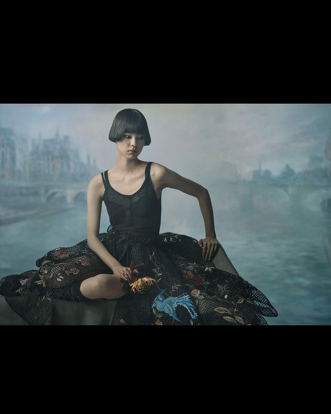 An adult female model sits in an embroidered, dark dress with dark hair in a bob with a blurred background that resembles a city.