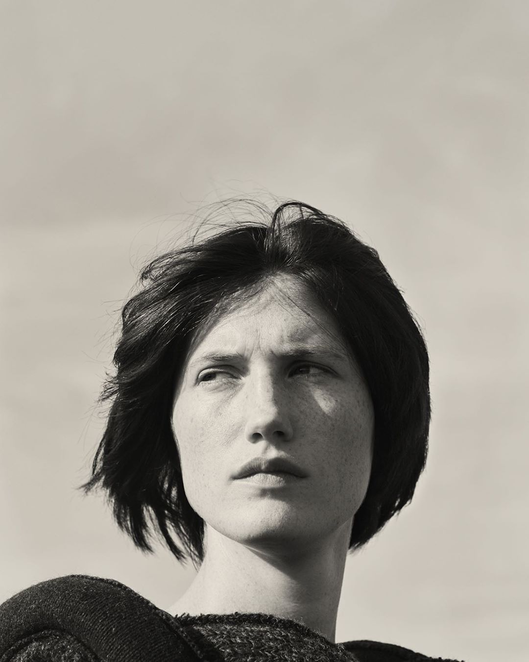 Monochromatic portrait of a fair-skinned person with short dark hair, looking off to the side, wearing a textured top against a sky background.