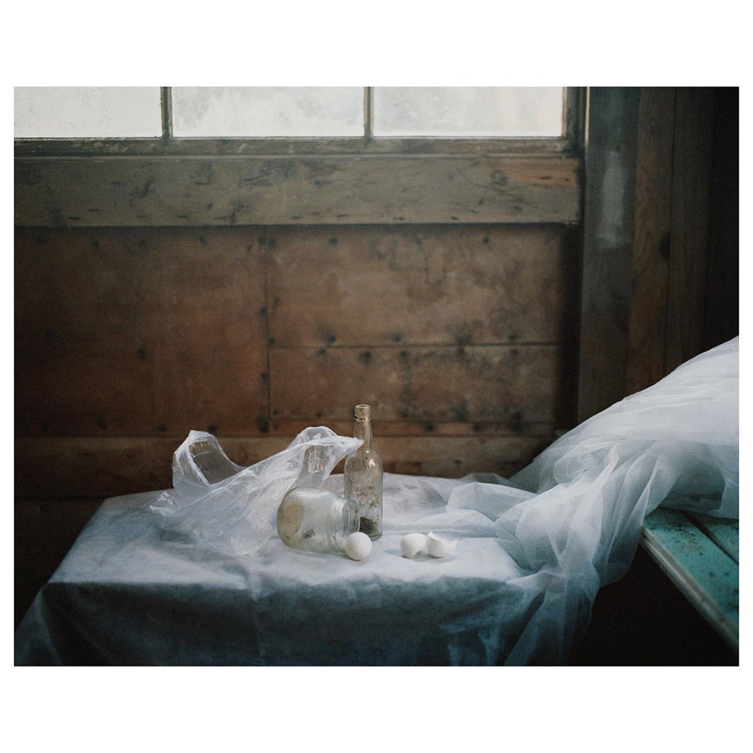 Still life photography featuring translucent plastic bag, glass bottles, and scattered eggs on a white tablecloth in a rustic room.