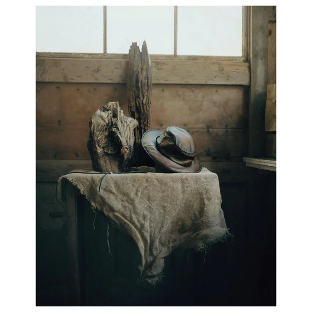 Still life photography of a vintage hat on a table with a textured cloth, alongside driftwood and a rustic wooden backdrop. 