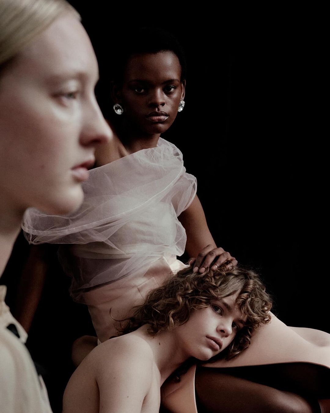 Three models pose in a studio environment, wearing sheer white and beige clothing, captured in a fashion-style portrait with a dark background.