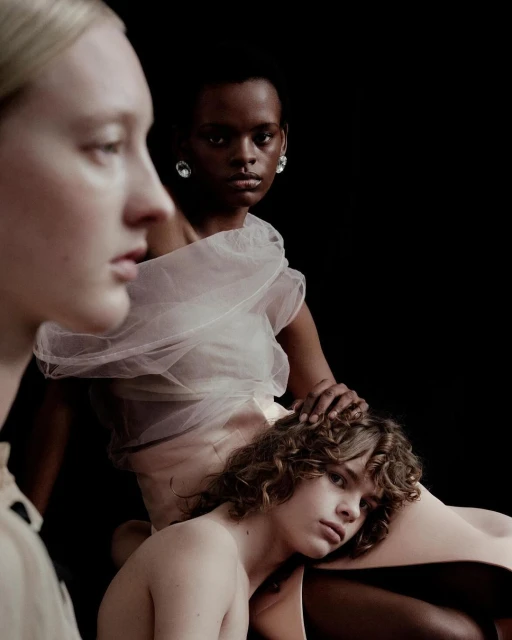 Three models pose in a studio environment, wearing sheer white and beige clothing, captured in a fashion-style portrait with a dark background.