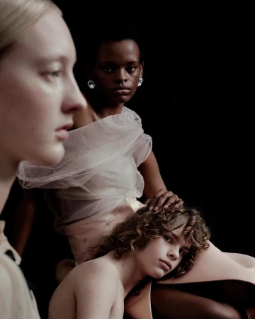 Three models pose in a studio environment, wearing sheer white and beige clothing, captured in a fashion-style portrait with a dark background.
