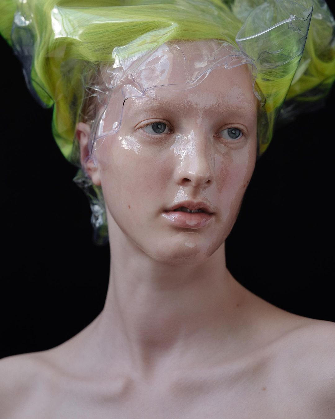 Close-up portrait of an adult woman with light skin and blue eyes, with green and clear plastic head covering against a black background.