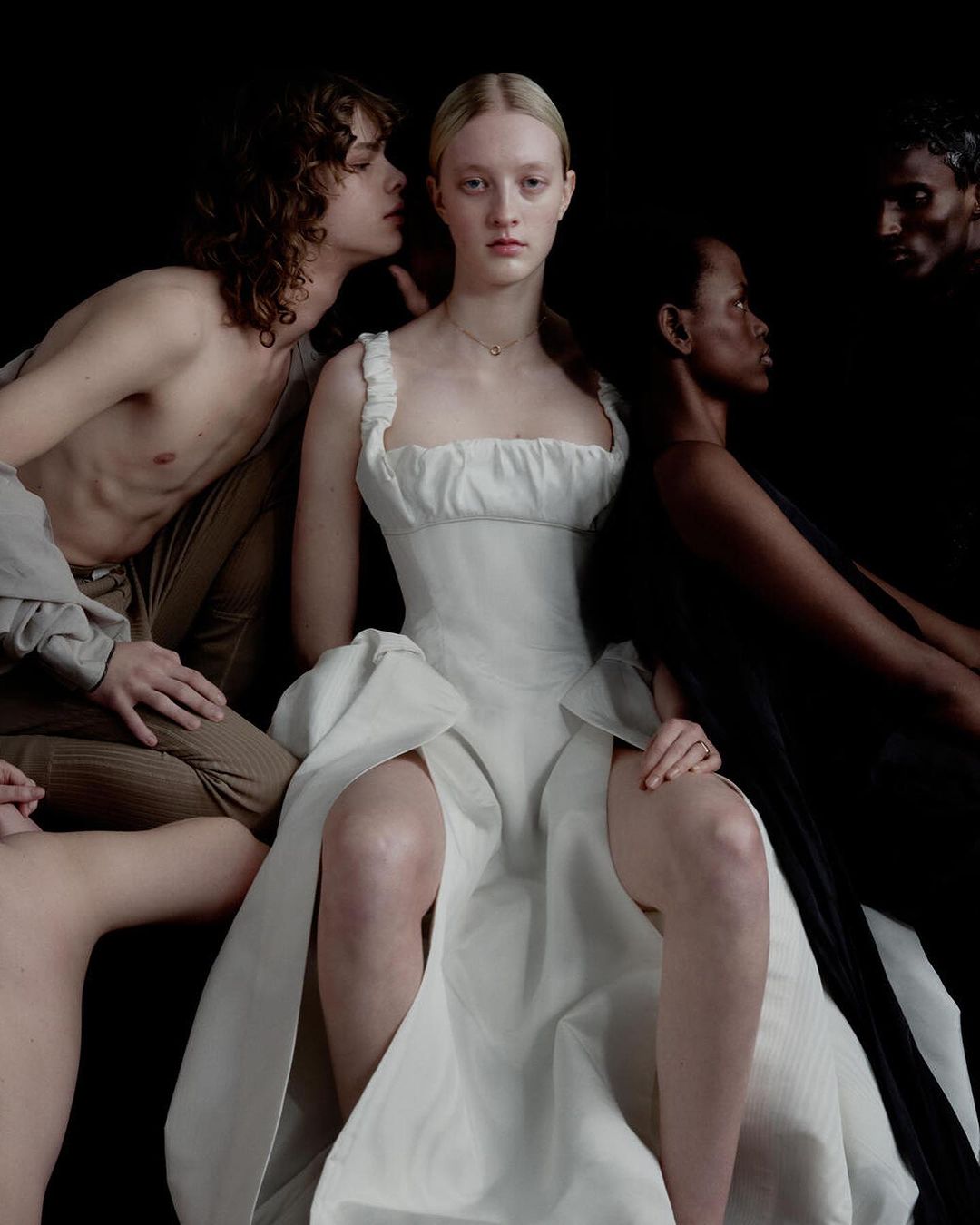 Group of four models posing in dark atmospheric lighting, centered on a woman in a white dress