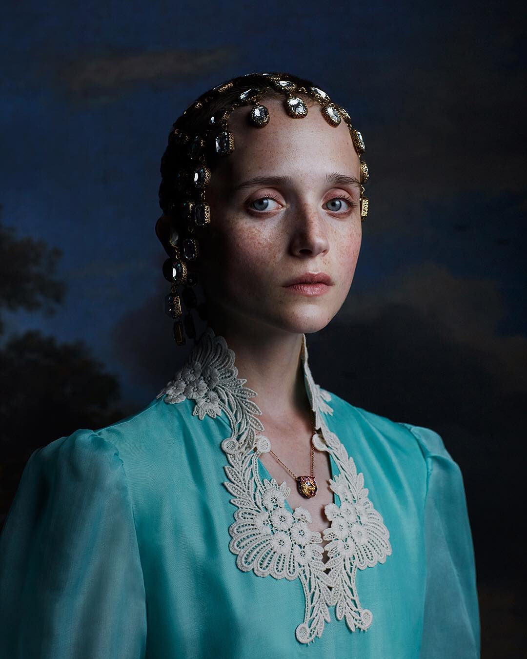 Portrait of a young woman wearing a turquoise gown with lace and an ornate crystal headpiece against a dark cloudy backdrop.