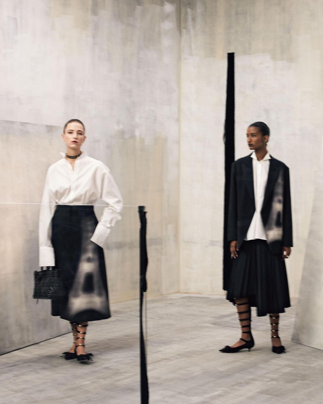 Two fashion models in elegant black and white attire pose in a minimalist photo studio with soft, diffused lighting and a stylish backdrop.