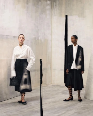 Two fashion models posing in an interior space wearing white shirts and dark skirts with patterned details