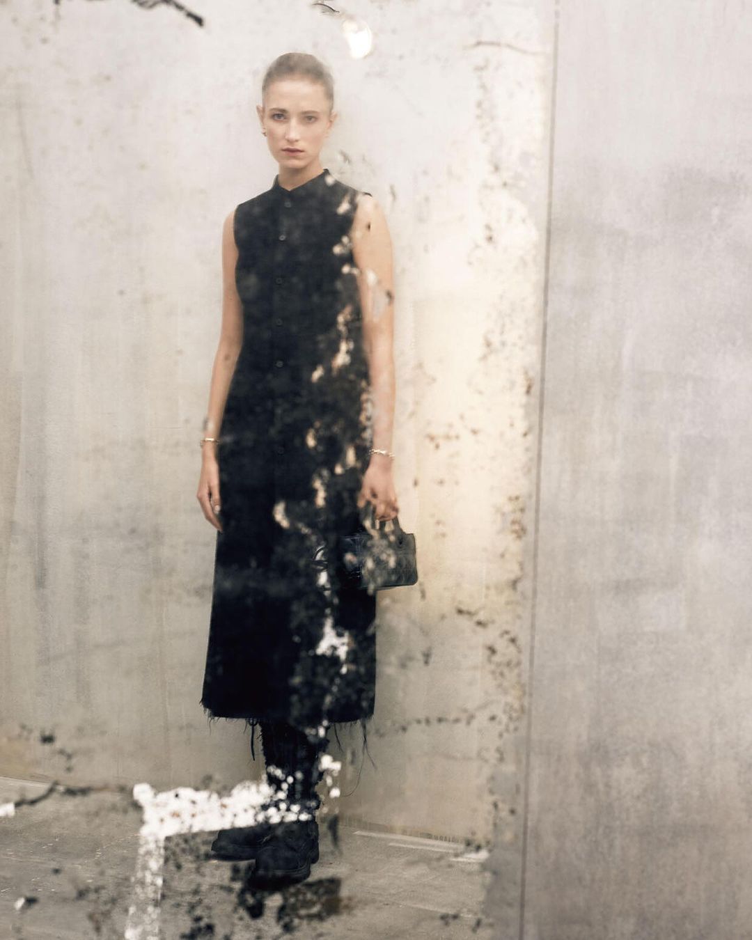A woman posing in a dark dress with a purse in an indoor setting against a concrete-style wall, with faded textures and light neutral tones.