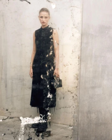 A woman posing in a dark dress with a purse in an indoor setting against a concrete-style wall, with faded textures and light neutral tones.