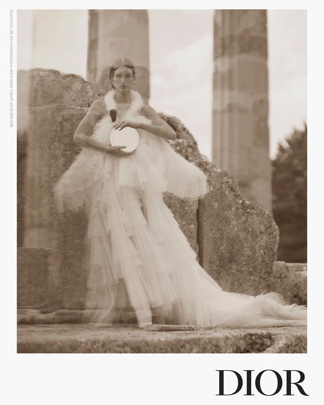 A woman in an elegant white dress poses near stone pillars, holding a round mirror. The image has a vintage, sepia-toned aesthetic.