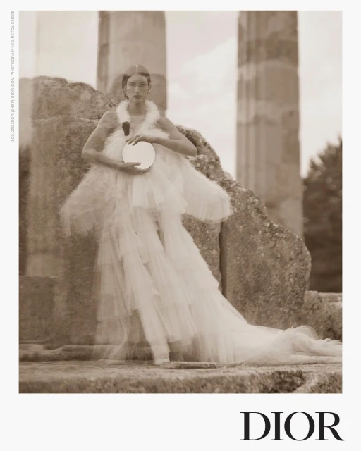 A woman in an elegant white dress poses near stone pillars, holding a round mirror. The image has a vintage, sepia-toned aesthetic.