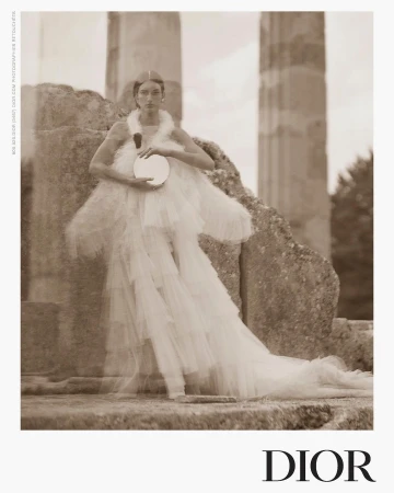A woman in an elegant white dress poses near stone pillars, holding a round mirror. The image has a vintage, sepia-toned aesthetic.