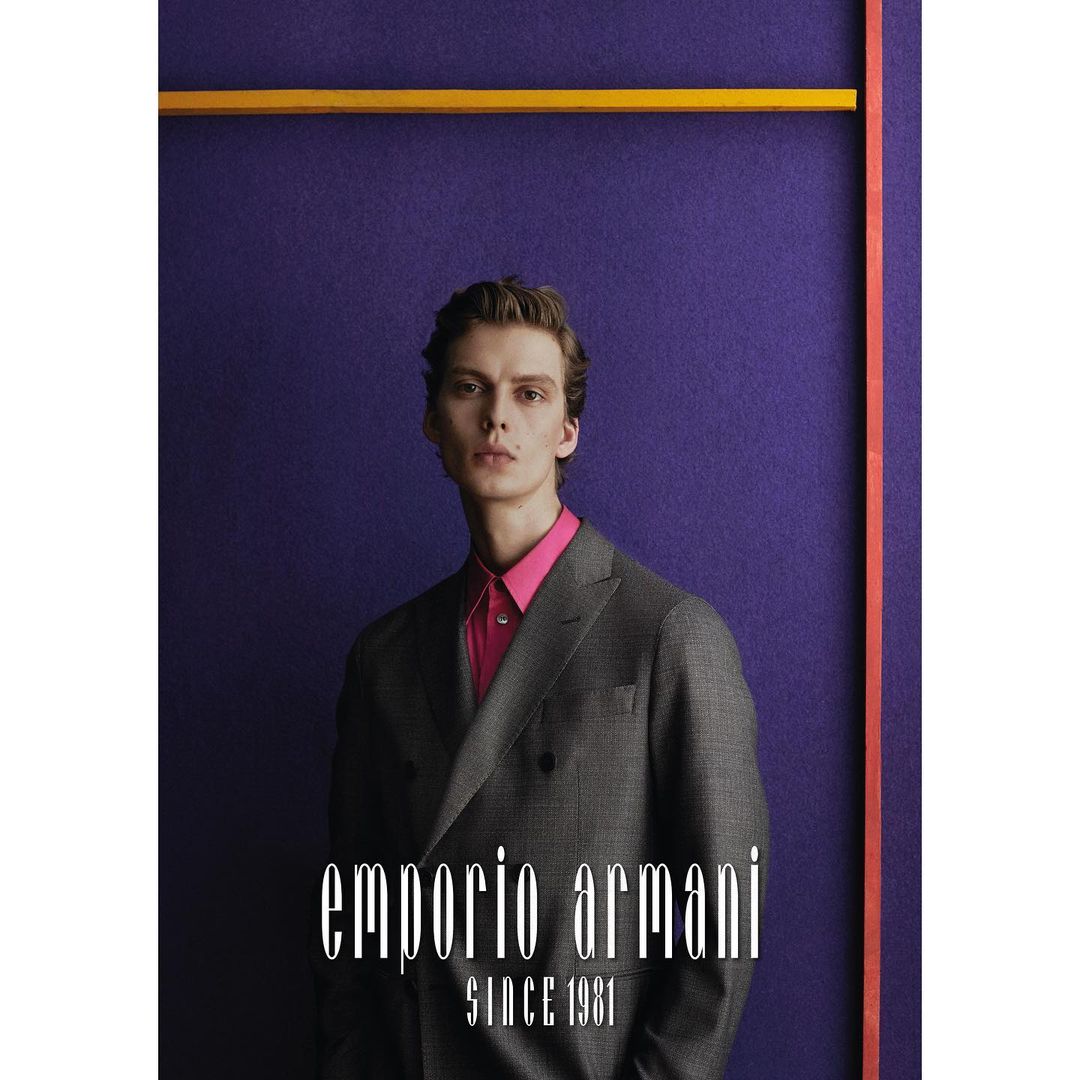 Man in a gray suit and pink shirt poses in front of a purple backdrop with a yellow and orange geometric accent; Emporio Armani logo.