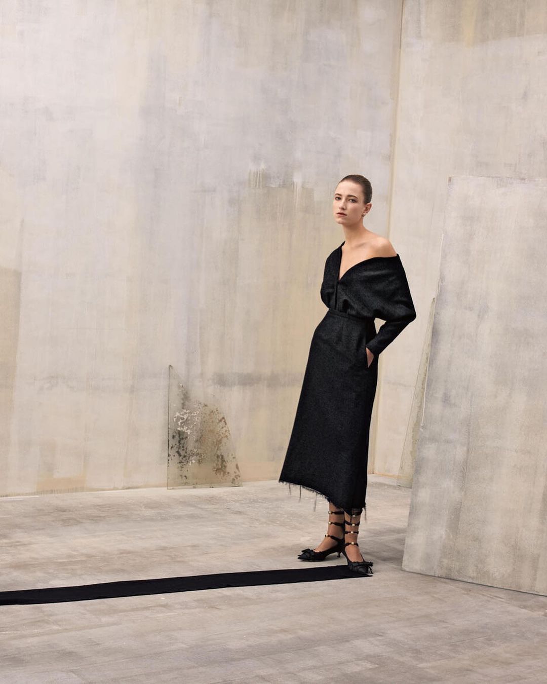 Fashion shoot: Woman in an off-shoulder black dress and black lace-up heels poses against a textured wall in a studio, looking elegant.