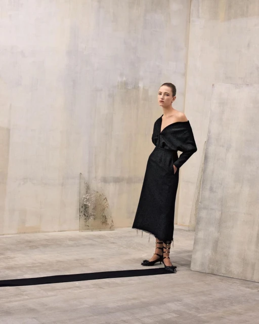 Fashion shoot: Woman in an off-shoulder black dress and black lace-up heels poses against a textured wall in a studio, looking elegant.
