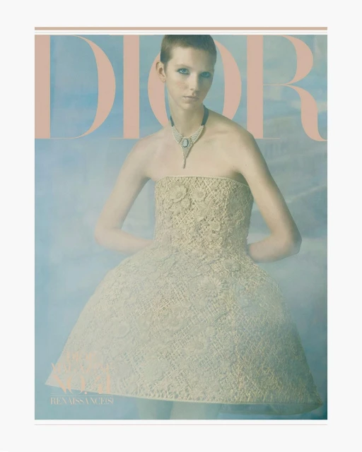 Fashion editorial portrait of a woman in a beige lace dress with a jewelry necklace, posing in front of a misty light blue background.