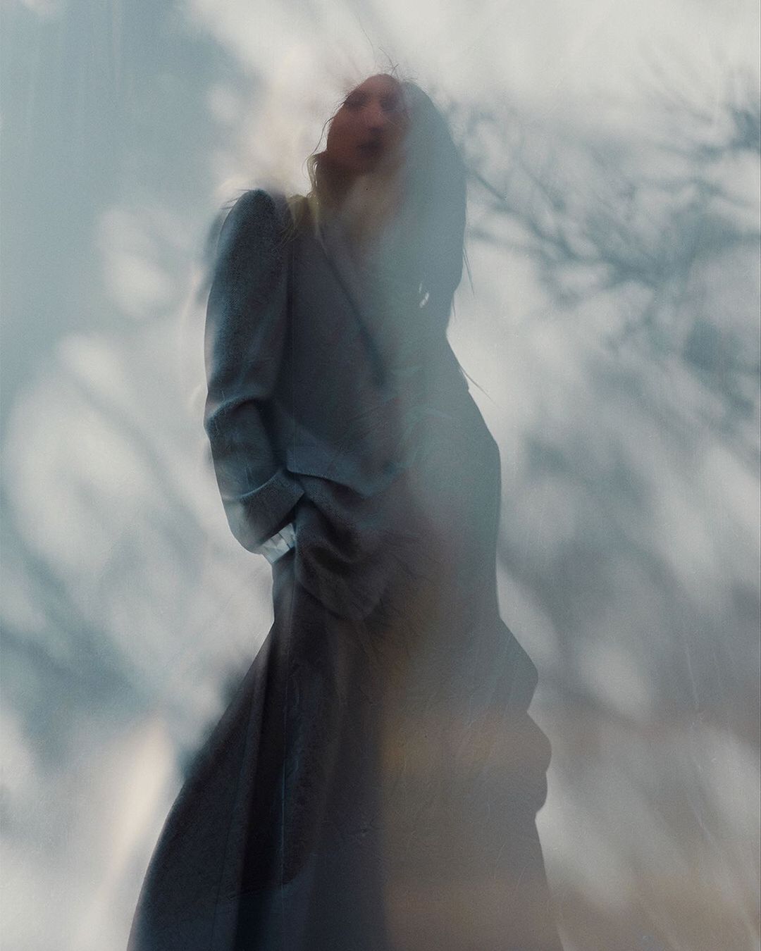 Model standing outdoors wearing a long grey coat viewed from a low angle through soft atmospheric blur.