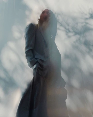 Model standing outdoors wearing a long grey coat viewed from a low angle through soft atmospheric blur.