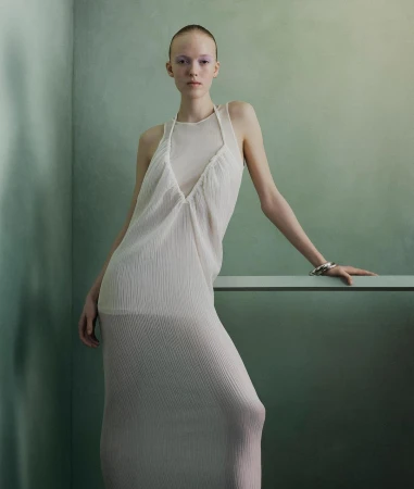 Fashion portrait of a fair-skinned adult woman posing in a long, light dress in front of a textured green backdrop with soft, diffused lighting.