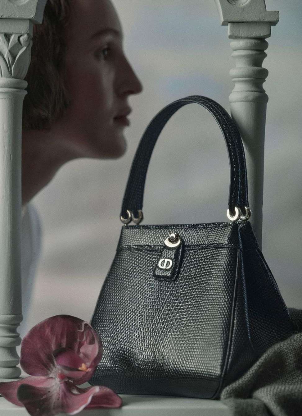 Fashion still life featuring a black textured leather handbag, set against a soft blurred backdrop with a model visible in the background.