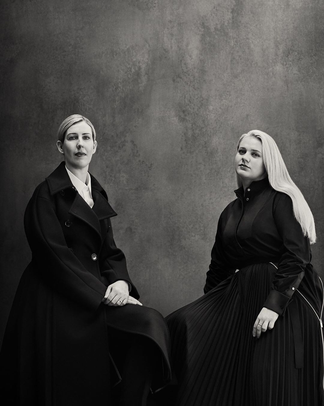 Two women in monochrome fashion pose in a photo studio. The first wears a coat, while the second dons a pleated skirt and dress shirt.