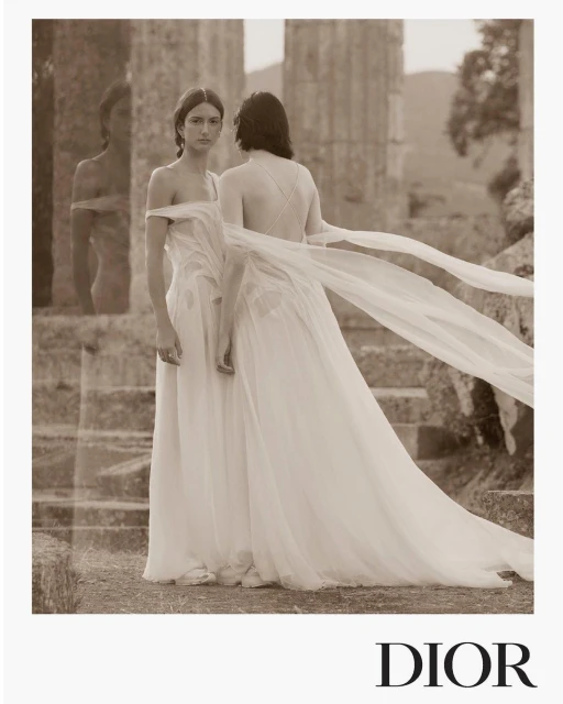 Two women in off-white gowns pose near stone ruins in a sepia-toned fashion shot for Dior, with long flowing fabric details.