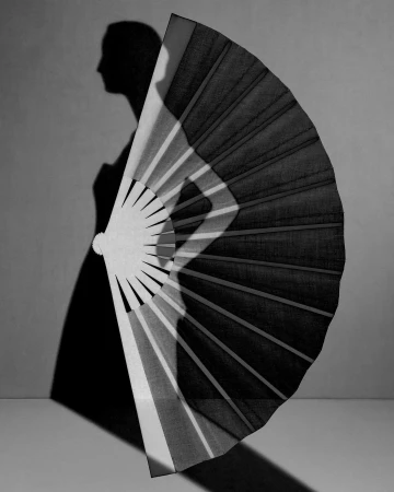 Monochrome image features woman's shadow behind a large, open fan, creating a dramatic interplay of light and dark against neutral backdrop.
