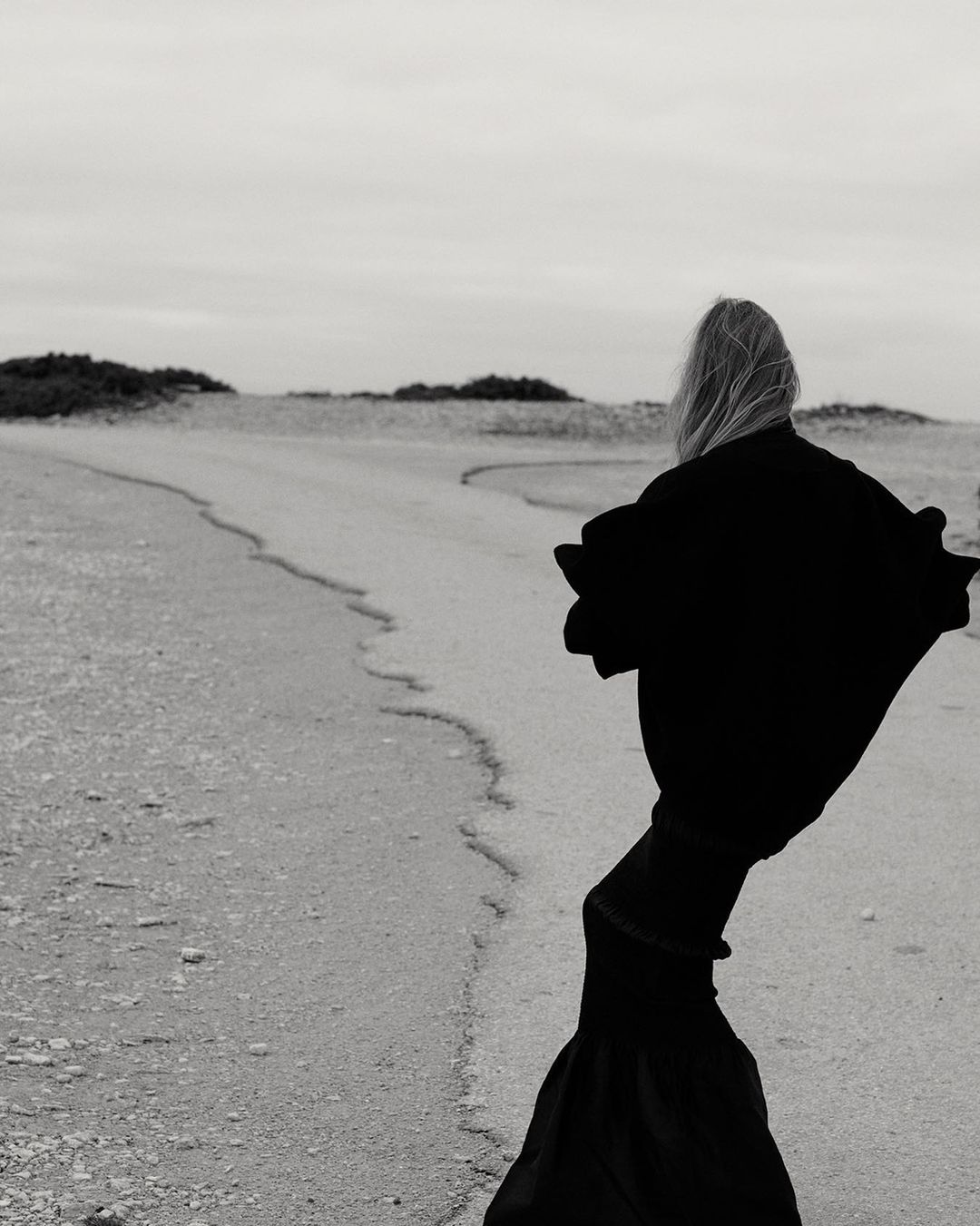 Person in dramatic black robe walks on sandy path towards horizon in black and white outdoor landscape under overcast sky.