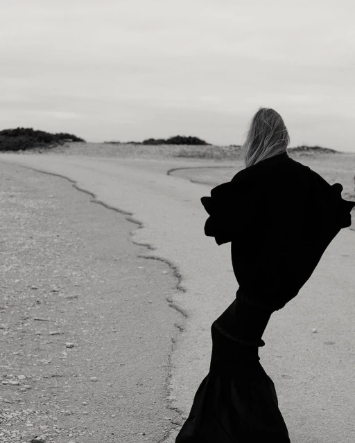 Person in dramatic black robe walks on sandy path towards horizon in black and white outdoor landscape under overcast sky.