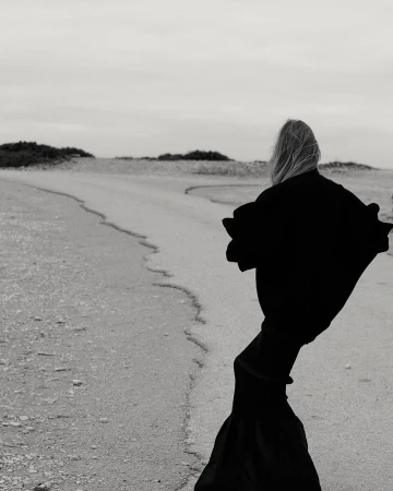 Person in dramatic black robe walks on sandy path towards horizon in black and white outdoor landscape under overcast sky.