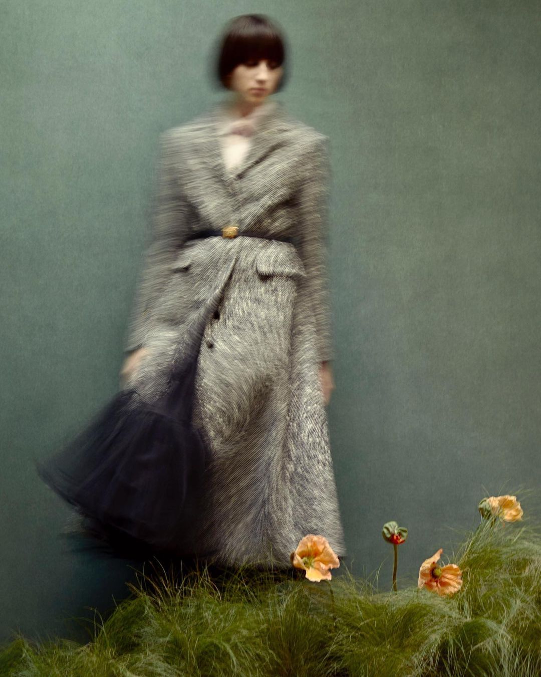 An adult female with short dark hair wearing a patterned gray coat and a black skirt posing in front of a green backdrop with flowers.