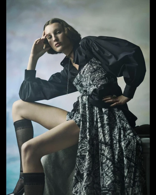 A young woman poses in a fashion shot, wearing a black top with a patterned skirt. She sits with her hand to her head, gazing forward.