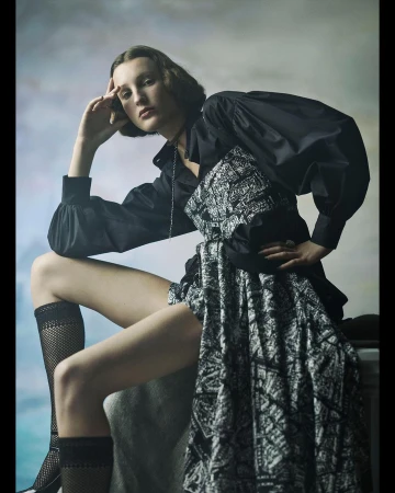 A young woman poses in a fashion shot, wearing a black top with a patterned skirt. She sits with her hand to her head, gazing forward.