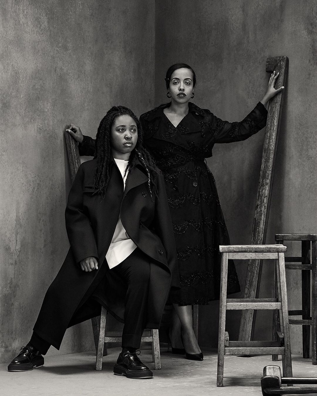 Two women pose in a monochrome portrait in a corner studio setting. One woman is seated on a stool, while the other stands next to a ladder.