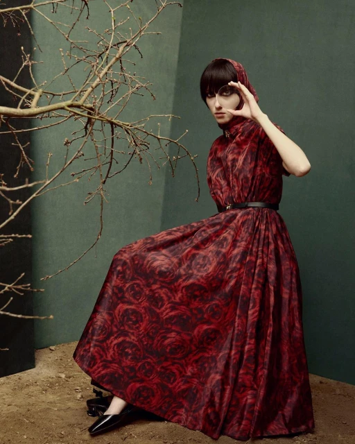Woman in a long red patterned dress sits in an indoor setting, framing her eye with her hand, with a stark branch to the left.