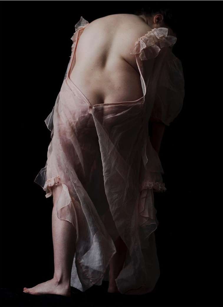 Back view of a slender woman in a pale pink negligee against a dark background, emphasizing her posture and the garment's texture.