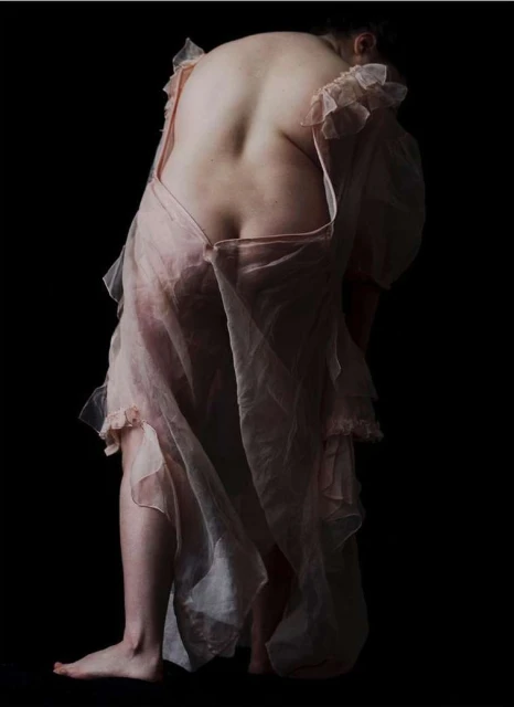Back view of a slender woman in a pale pink negligee against a dark background, emphasizing her posture and the garment's texture.