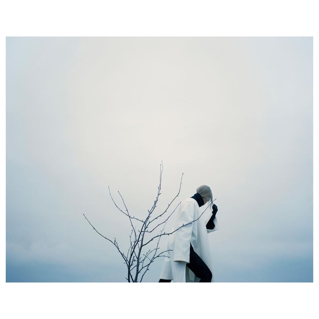 Person wearing a white coat with black accents standing near a bare tree against an overcast sky, creating a minimalist and eerie composition.