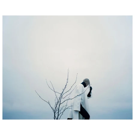 Person wearing a white coat with black accents standing near a bare tree against an overcast sky, creating a minimalist and eerie composition.