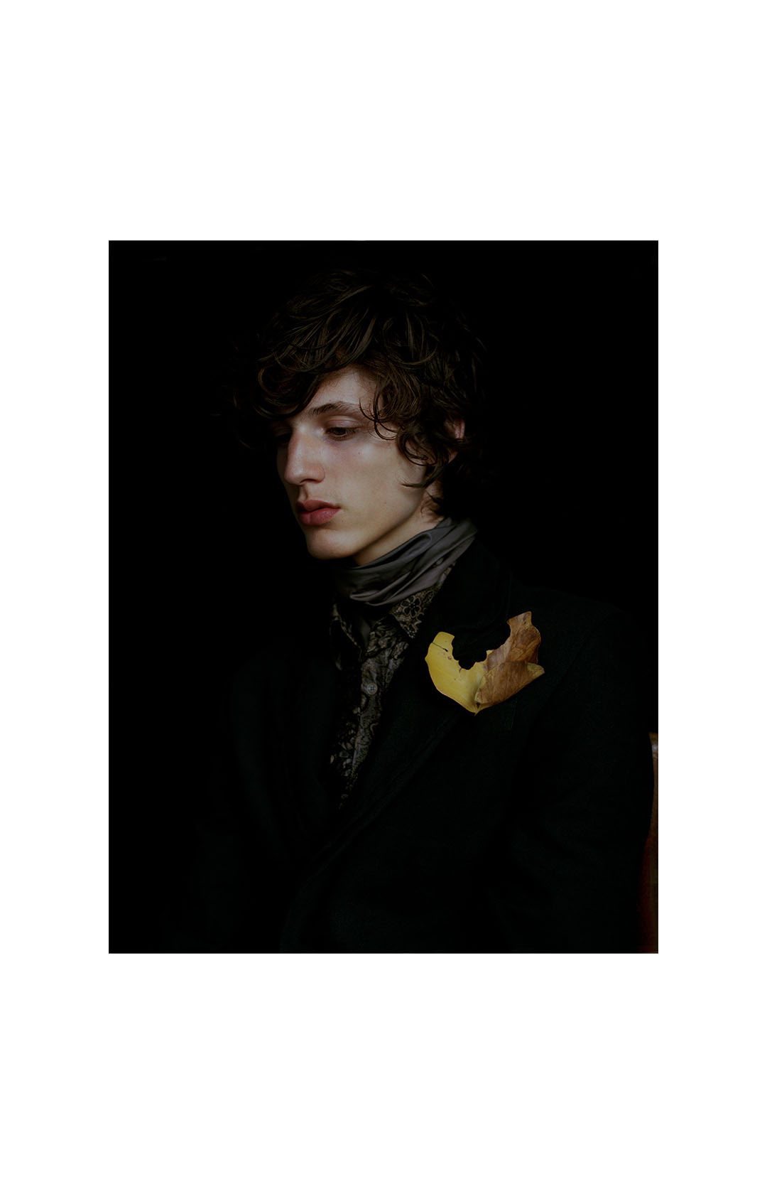 Portrait of a young adult man with dark hair wearing a jacket, a patterned shirt and a scarf, with a brown leaf attached.