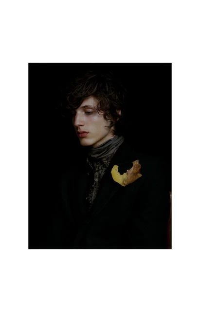Portrait of a young adult man with dark hair wearing a jacket, a patterned shirt and a scarf, with a brown leaf attached.