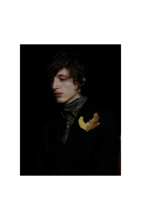 Portrait of a young adult man with dark hair wearing a jacket, a patterned shirt and a scarf, with a brown leaf attached.