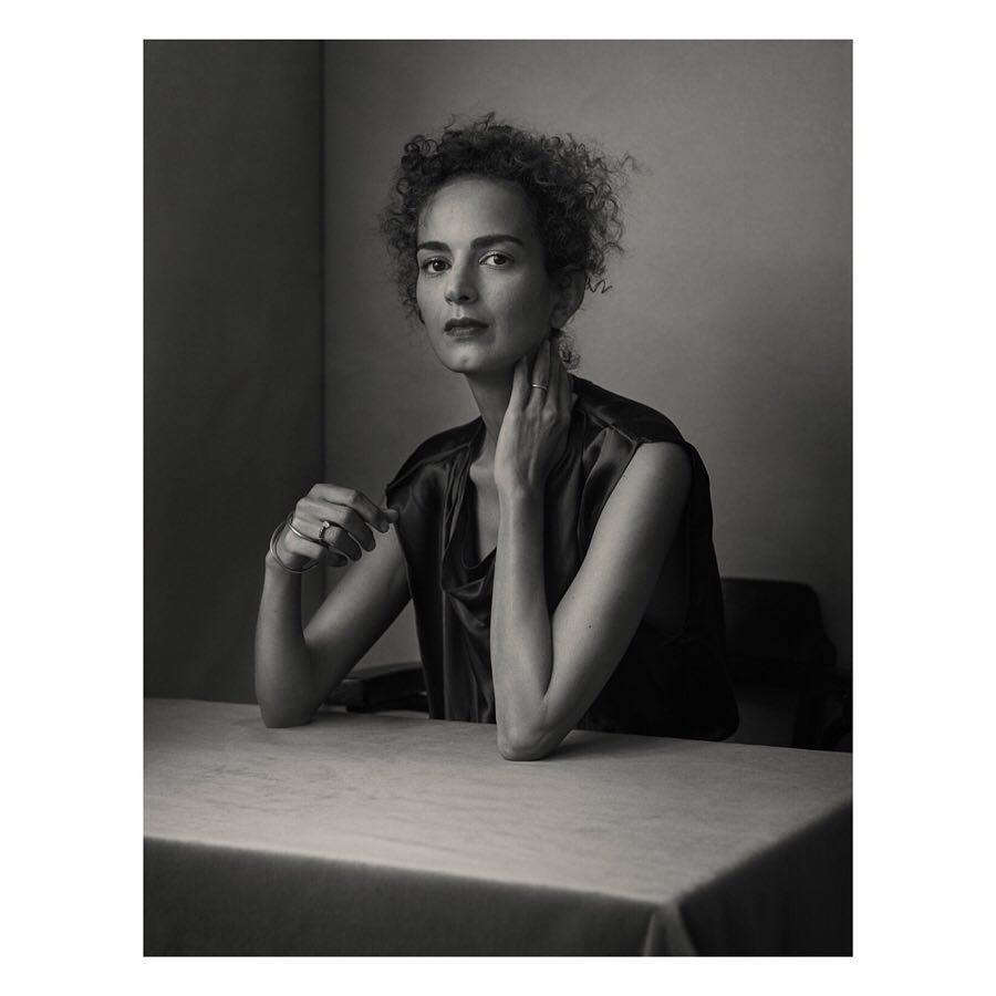 Monochrome portrait of a woman with curly hair seated at a table, captured in a photo studio with soft, diffused lighting.