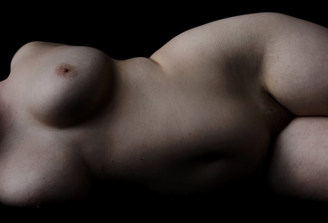 Nude torso of an adult female lying on her side in a dimly lit black studio with a full view of her breast and midsection.